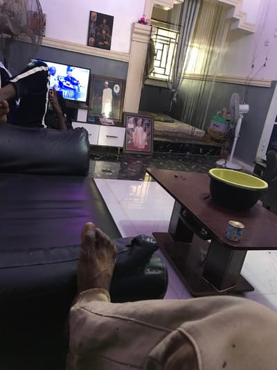 A room contains a sofa and a desk with a speaker, along with a can of soda. A person is sitting on the sofa watching TV. Several framed pictures are hanging on the wall. One person's bare foot is resting on the arm of the sofa. The room is lit with both natural and artificial light. A ceiling fan and a standing fan are present.