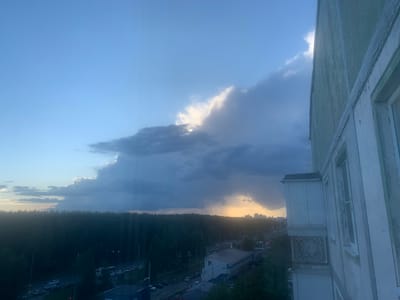 This image depicts an outdoor scene in Ivanovo, Russia, likely during the late afternoon or early evening, as indicated by the soft, golden light breaking through dramatic clouds. The sky is a dominant feature, with a vast expanse of blue sky partially obscured by large, dark storm clouds that are beginning to break, allowing sunlight to stream through. A patch of lighter, illuminated clouds suggests rain might be occurring in the distance.
In the foreground, the side of a multi-story building is visible, its light green facade showing signs of age and weathering. The building's architectural details, such as window frames and decorative elements, are partially visible. Below the building, a dense line of dark green trees forms a tree line, suggesting a natural or park-like area bordering the urban landscape.
Beyond the trees, a street is visible with several cars and what appear to be buses. This indicates ongoing traffic and a populated area. In the far distance, a city skyline with several buildings is faintly discernible, silhouetted against the illuminated sky. The overall atmosphere is one of dramatic weather, with the contrast between the dark clouds and the emerging sunlight creating a sense of anticipation or transition. No people are clearly visible, and no text is legible in the image.