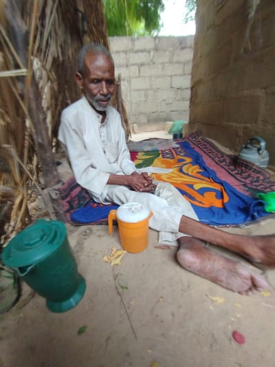 An elderly man with a white beard is sitting on a colorful blanket in a sandy outdoor area,  in Bajoga, Nigeria (coordinates: 10.851, 11.446).  He is wearing a long-sleeved white shirt and white pants and has his legs extended in front of him. He is resting his hands on his lap. There is a green water jug beside him and an orange water jug next to his legs. The blanket is on the ground, and the wall of a building is to the right, partially obscured by the blanket. Behind the man is a wall made of brick. The area is sparsely vegetated.  There is a green object in the foreground that appears to be a plastic container. The ground is sandy.  The overall scene is one of a man relaxing on a blanket outdoors.