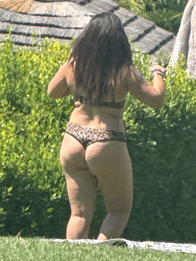 A woman, viewed from the back, stands on grass in front of dense green foliage. She wears a leopard-print bikini. Her hair is dark and long, reaching down her back. She has a bracelet on her left wrist. Behind her, a thatched roof structure is visible, and to the right, a portion of a pale, textured object, possibly a tree trunk or a pillar, is present. The scene appears to be outdoors on a sunny day.