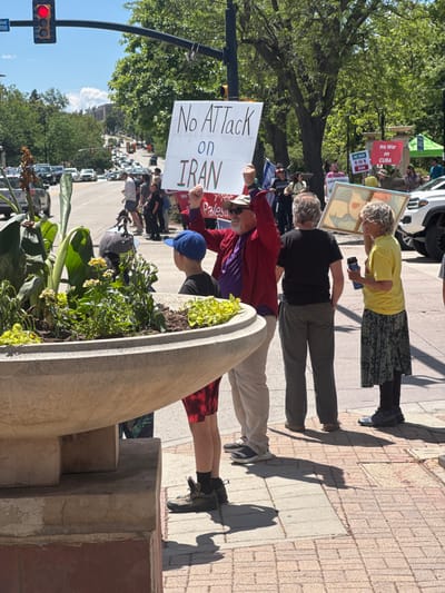 The image shows a political demonstration in Boulder, Colorado. Several people are standing on a sidewalk holding signs that protest potential military action against Iran and Cuba. One sign reads "No Attack on IRAN." Another states "No War on CUBA." A third reads, "Palestine." The protesters appear to be on a street with a considerable amount of car traffic and trees alongside the sidewalk. There is a traffic light visible in the bottom left corner of the frame, and a sign on the traffic light that reads "Blvd 1700."
