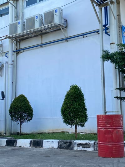 In Gambut Barat, Indonesia, a red metal barrel stands to the right of the frame, positioned in front of a white building. Three outdoor air conditioning units are mounted on a metal rack attached to the building's exterior wall. Two small, cone-shaped green trees are planted in front of the building, with a grassy area behind a curb separating them from the paved ground. Blue pipes are visible running along the building's exterior. The scene appears to be an outdoor industrial or commercial area.