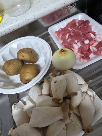 The image displays an overhead view of food ingredients arranged on a kitchen counter, likely in a residential setting in Daejeon, South Korea. The ingredients include raw pork, potatoes, an onion, and king oyster mushrooms. The pork is thinly sliced and piled in a white styrofoam container, some pieces showing marbling. Three whole potatoes are in a white oval bowl. A single, pale yellow onion sits next to the pork. A large pile of king oyster mushrooms, with their characteristic thick stems and flat caps, are arranged on another white styrofoam container. The countertop appears to be a speckled, light-colored material, and a stainless steel sink is visible in the background. The lighting suggests an indoor environment, possibly a home kitchen. There are no people or actions depicted in the image.