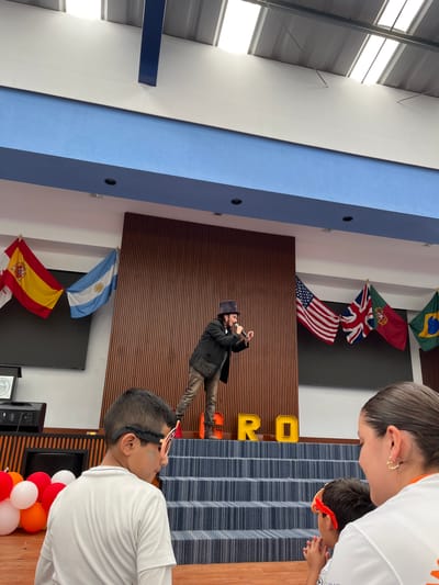 The image depicts an indoor scene, possibly a school or community center, in Tlaxcalancingo, Mexico. The walls are adorned with flags from various countries, including Brazil, Portugal, the United Kingdom, the United States, Argentina, and Spain. A man in a dark suit and hat stands in front of a brown-striped wall, seemingly taking a photo with his phone. In the foreground, people wearing white shirts are visible, including children with party accessories. Balloons in red, white, and orange colors are near the bottom right.