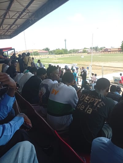 The image shows a view from the stands of a sports field in Isasi, Nigeria. The field is visible in the background, surrounded by a low fence and some buildings. In the foreground, a crowd of people are seated in the stands. The people are mostly young men, and they are wearing a variety of clothing. Some of the people are looking at the field, while others are talking to each other. The stands are covered by a metal roof.