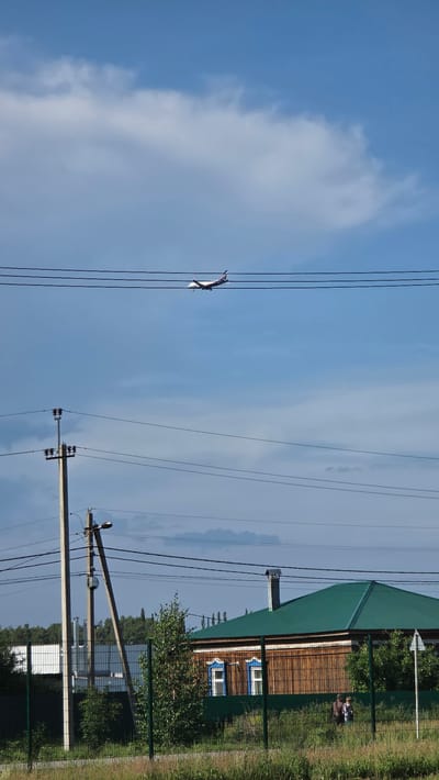 An airplane is seen flying in the sky above a residential area with a wooden house, power lines, and a fence in Bulgakovo, Russia. The sky is partly cloudy, with a mixture of blue sky and white clouds. The airplane is a commercial jet with a white fuselage and a blue and red tail. It is flying at a moderate altitude. The house is a single-story wooden structure with a green metal roof. The windows have decorative blue trim. Two people are visible near the house, standing behind a green metal fence. The area is surrounded by trees and grass. Power lines run across the foreground and middle ground of the image, supported by concrete poles.