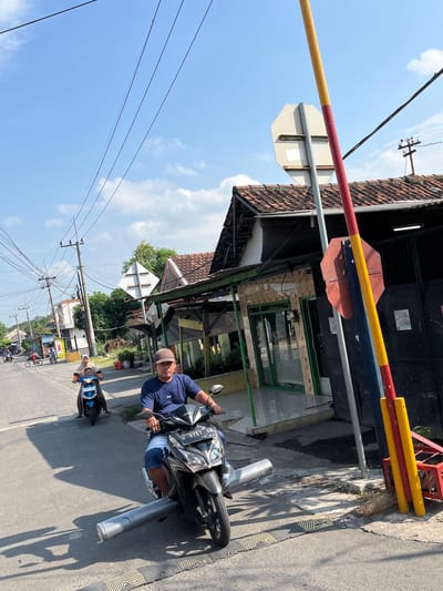 A man wearing a blue t-shirt and shorts rides a black motorcycle down a paved street in Kludan, Indonesia, under a clear blue sky with scattered clouds. The motorcycle has a license plate that reads "W 2757 OB". In front of the motorcycle, there is a speed bump, and the motorcycle is carrying a long, cylindrical object wrapped in silver material. To the left of the man, a woman wearing a headscarf rides a blue motorcycle with a child behind her. In the background, there are several houses with tiled roofs and utility poles with electrical wires. A red and yellow striped pole, possibly part of a railroad crossing, stands on the right side of the frame.