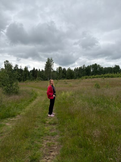 A blonde woman stands on a dirt path in a grassy field, looking over her left shoulder towards the camera. She is wearing a red jacket, black pants, and pink athletic shoes. Behind her, the field extends to a dense line of trees under a cloudy sky. The location is a field in Vaskelovo, Russia.