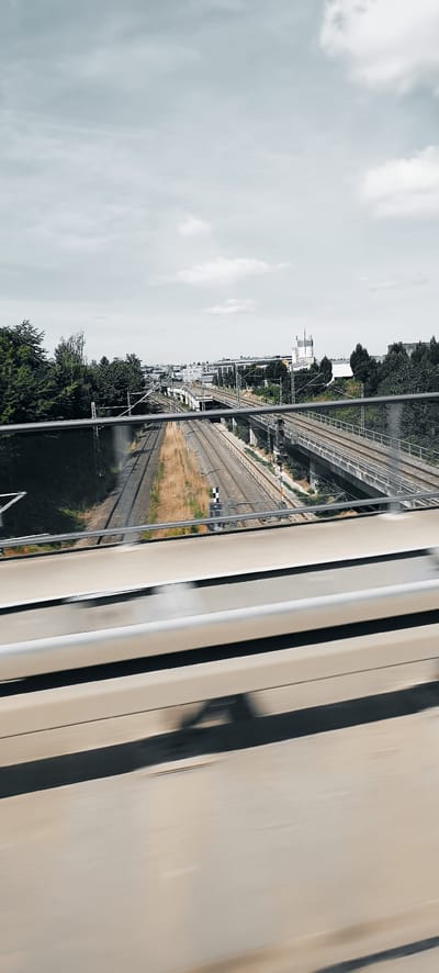 The image shows a view from a bridge overlooking multiple train tracks in Waiblingen, Germany. The sky is overcast with clouds. In the foreground, a metal guardrail runs horizontally across the frame. Below the guardrail, blurred motion streaks indicate the perspective is from a moving vehicle.

Further down, several sets of train tracks run parallel to each other. Some tracks are elevated on concrete structures, while others are at ground level. On the left side of the train tracks, dense green trees line the embankment. On the right side, there are more elevated train tracks with some greenery beneath them. In the background, there are buildings and structures consistent with an urban or industrial area. A railway signal with a black and white checkered pattern is visible next to the ground-level tracks. The overall scene depicts a transportation hub with extensive railway infrastructure.