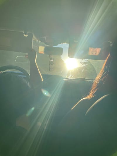 Inside a vehicle, glare from the sun obscures much of the scene. A driver's arm rests near the steering wheel. The sun shines through the windshield, partially blocked by a sun visor displaying an image of a mouth. A side mirror is visible. The dashboard and parts of the vehicle's interior are discernible amidst the bright sunlight and shadows. Some hair is visible at the top of the frame.