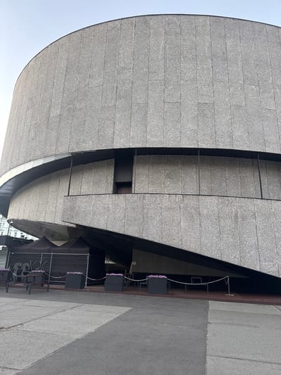 The image shows a section of a building's exterior with a curved, grey, textured facade made of rectangular panels. The building's design features sharp angles and overhangs, creating a modern architectural style. Below the overhang, there is an outdoor seating area with black tables and chairs, some of which are stacked. Two black planters filled with purple flowers are placed in front of the seating area, with decorative fencing surrounding it. The ground is paved with grey concrete tiles and asphalt. The sky is a pale, overcast grey, suggesting it might be daytime with cloudy weather. The scene appears to be located in an urban or public space, possibly a cafe or restaurant with outdoor seating. There are no people visible in the image, and no text is discernible. The image's orientation is rotated 90 degrees counter-clockwise.