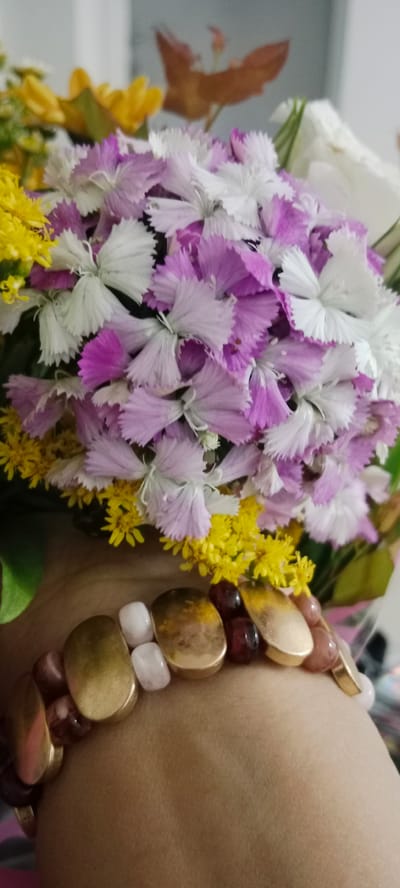 A close-up, high-angle shot captures a bouquet of flowers with a bracelet worn on a person's wrist. The bouquet contains numerous small, delicate flowers, predominantly in shades of purple and white, interspersed with clusters of small yellow flowers. A few green leaves are visible at the edges of the bouquet. The bracelet features a mix of beads, including oval, metallic gold-toned pieces, translucent pinkish beads, and darker reddish-brown beads. The background is blurred, suggesting an indoor setting, possibly a home environment in Bnei Brak, Israel. The lighting is soft and diffused.