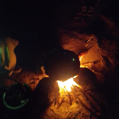 The image depicts a nighttime scene, likely outdoors, with a campfire as the central element. The fire is a bright, yellowish-orange, illuminating the surrounding area. To the left of the fire, a dark, amorphous object with some green and orange hues is visible; it appears to be a pot or container with some contents. Further to the left, an object with a curved, orange shape is present, possibly a piece of equipment or furniture. In the background, several dark shapes suggest the presence of people, with a pair of feet wearing sandals visible at the top right. The ground appears to be sandy or earthy, with some rocks and embers scattered around the fire. The overall ambiance is one of a gathering or camping at night. The location in the city of Shuwa, Nigeria, cannot be determined from the visual information provided in the image.