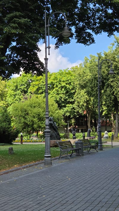The image shows a paved walkway in Palanga, Lithuania, leading into a park. Two ornate, dark grey lampposts stand on either side of the walkway, with one in the foreground to the left and another further back to the right. In the middle ground, there are two dark green park benches facing the walkway. Behind the benches, a large, dark sculpture is visible, appearing to depict several figures. Further into the park, there is a patch of grass with scattered small white flowers, and behind that, a line of yellow flowers. Numerous trees with lush green foliage form the backdrop, with parts of buildings peeking through the trees on the right side of the image. The sky above is blue with scattered white clouds. The overall scene is one of a peaceful, well-maintained public park in Palanga.
