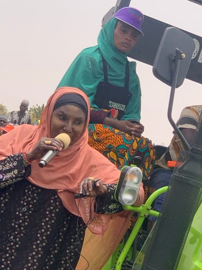 The image shows a woman standing on a small tractor, wearing a pink hijab and a black and white dress, speaking into a microphone. She is holding the microphone in her right hand and appears to be giving a speech. Behind her, a second woman wearing a green headscarf with an orange and yellow skirt sits in the driver's seat. She is wearing a black apron with "WORN" printed on it and a purple cap. A third person is in the back of the tractor, partially obscured by the frame, wearing a light colored shirt. The scene takes place in Ringimawa, Nigeria. The tractor is a small green vehicle with a large black head lamp. The background consists of a blurred view of the surrounding area.