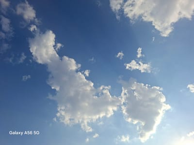 A bright blue sky is filled with scattered white cumulus clouds. Sunlight streams from the upper right, creating a bright halo and illuminating the clouds from behind. The lower left corner of the image displays the text "Galaxy A56 5G" in white, indicating it is a photograph taken with a Samsung Galaxy A56 5G phone. No specific location within the city of Obshoron, Tajikistan can be determined from this image, as it solely depicts a sky scene.