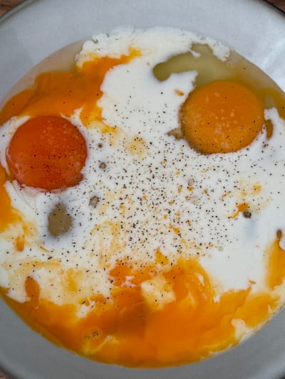 A top-down view of a light gray bowl containing two raw egg yolks, some egg whites, and a white liquid, possibly milk or cream. Black pepper is sprinkled over the mixture. The bowl appears to be sitting on a wooden surface. No information is available relating to Bjeliši, Montenegro.