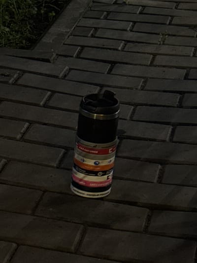 A black thermal mug with a silver band around the top rim stands vertically on a brick pathway. The mug is adorned with several horizontal bands of colorful stickers, each featuring various logos and text, suggesting travel or event participation. The pathway is made of dark, rectangular bricks laid in a herringbone pattern. To the left of the mug, a patch of green grass with some sparse vegetation is visible, bordered by a raised concrete edge. The overall lighting suggests it is either dusk or night, with shadows cast on the brickwork. The location is Doma 1096 Km, Russia.
