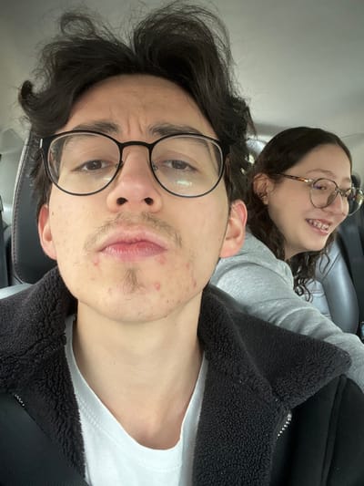 A close-up photograph captures two individuals inside a vehicle, located within the city of Bogota, Capital District, Colombia. The primary subject in the foreground is a young male, facing the camera with a neutral expression and slightly pursed lips. He has dark, voluminous hair, wears round, black-framed eyeglasses, and displays a sparse mustache and goatee with visible acne on his chin and lower cheeks. He is dressed in a white t-shirt beneath a black jacket featuring a fleece-lined collar. Behind him, to the right and slightly out of focus, a young female is visible. She has dark, wavy hair, wears light-colored, round-rimmed eyeglasses, and exhibits dental braces on her upper teeth. Her head is turned to her right, away from the camera, and she is smiling. She is wearing a light grey hooded sweatshirt. The interior environment shows dark vehicle seat upholstery and headrests.