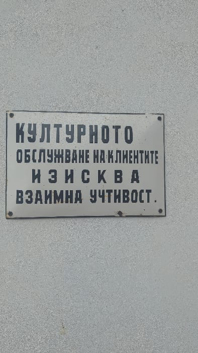 A rectangular, light grey metal sign with black Cyrillic text is affixed to a textured, light grey wall in Slokoshtitsa, Bulgaria. The sign is secured by four dark rivets, one in each corner, with minor corrosion visible around the bottom two and along the bottom edge. The text, translated from Bulgarian, reads: "Cultural service to clients requires mutual courtesy." The wall surface exhibits a fine granular texture consistent with stucco or rough concrete. No individuals or dynamic actions are present in the frame.