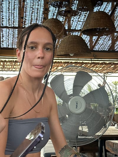 A woman is featured in a close-up, rotated portrait, appearing to take a selfie or be photographed in what looks like a cafe, restaurant, or covered outdoor area in Hà Giang 1 Ward, Vietnam. She has dark hair styled in several braids, a light complexion, and is wearing a dark grey strapless top and a thin gold chain necklace. Her expression is playful, with her tongue slightly extended.
To her right, a white Hatari brand standing or ceiling fan is prominent, its blades visible behind a protective grille. Multiple woven, basket-like light fixtures or decorative elements hang from the ceiling in the background. The setting features light filtering through a patterned, possibly bamboo or rattan, ceiling or wall structure, suggesting a rustic or tropical aesthetic. Bright natural light illuminates the scene, indicating it's daytime. A tattooed arm is visible on the right, holding a reflective object, likely a smartphone. The overall impression is casual and relaxed.