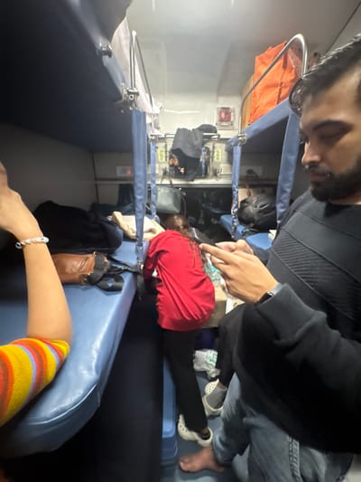 A crowded train compartment shows multiple passengers in close quarters. A man in a black sweater stands above a woman in a red shirt who is lying down on a blue seat. Another person sits in the foreground, wearing a striped sock and a bracelet. Luggage is scattered around, indicating a long journey. The lighting is dim, casting shadows and creating a sense of shared, intimate space.