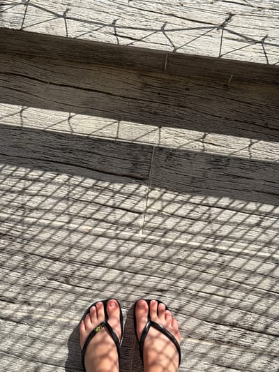A high-angle, close-up photograph displays two human feet, likely adult, wearing black flip-flops, positioned on a textured, light grey-brown plank surface. The strap of the left flip-flop features a small emblem resembling the Brazilian flag, consisting of green and yellow colors. The surface, appearing to be weathered wood or a wood-mimicking material, is extensively covered by strong, diagonally oriented grid-patterned shadows, cast by an unseen overhead or adjacent lattice-like structure. This shadow pattern indicates direct sunlight illumination. The scene is situated outdoors in Porto Alegre, Brazil; however, a specific address or landmark within the city cannot be determined solely from the visual information presented.