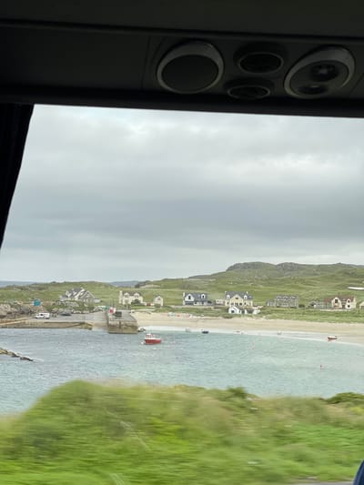 This media file depicts a picturesque coastal scene at Lagnalore, Ireland. The image is oriented vertically, with the left side dominated by a dark, textured surface that appears to be part of a vehicle's interior, possibly a window frame or dashboard, featuring several circular elements.

The main subject is a scenic view of a bay with a sandy beach, turquoise water, and a backdrop of grassy hills dotted with houses. The houses are varied in style, with some appearing traditional and others more modern. A small pier or jetty extends into the water, with a red boat moored alongside it. Several other small boats and buoys are visible scattered across the water.

The beach has a few scattered people, suggesting a leisurely day, and there are a few parked cars visible along the road near the waterfront. The sky is overcast, indicating a cloudy or possibly humid day. The overall impression is one of a serene, rural coastal settlement, likely in the summertime given the presence of people enjoying the beach and water. No visible text is present in the image.
