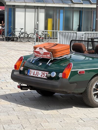 This media file shows the rear of a vintage green MG MGB convertible parked on a cobblestone street in Bruges, Belgium. The car is equipped with a luggage rack holding two vintage brown suitcases. A red sign attached to the rack reads "EDITIE 2021 BRU WEST BRUGGE CLASSIC TOUR". The license plate of the car is visible and reads "0 AJU - 257". In the background, a modern building with large glass windows and a bicycle rack with several bicycles can be seen. The lighting suggests it is daytime, and the shadows indicate a sunny day. The overall scene evokes a sense of classic car touring and exploration in a historic European city.
