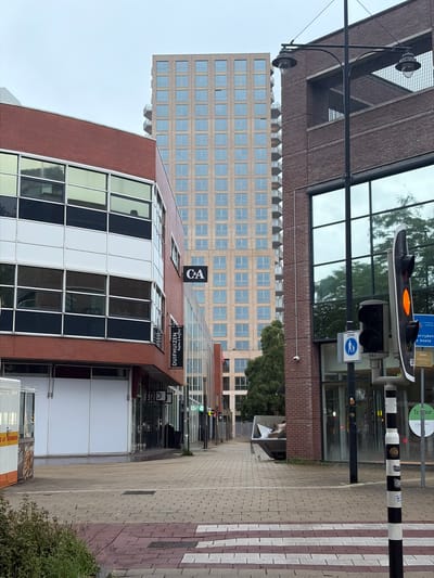 This image depicts an urban street scene in Rijswijk, Netherlands, specifically at the Generaal Vetterstraat within the "In de Bogaard" shopping center, facing the Rijswijkseplein residential tower. The view is from a paved pedestrian area on an overcast day.
In the background, a tall, modern residential tower dominates the skyline, featuring a light-colored facade with numerous uniformly sized rectangular windows. Its right side reveals several stacked balconies with metal railings.
In the mid-ground, on the left, stands a multi-story commercial building with a red brick base and a facade composed of large reflective windows interspersed with dark and white panels. A black "C&A" sign is mounted on its upper section, and a vertical black banner sign below it reads "DUIFHUIZEN Tassen & koffers". The lower-level windows appear obscured internally with white material.
On the right, another multi-story commercial building with a dark brown brick facade features large reflective windows that mirror nearby trees and the sky. An elevated, recessed section with dark metal railings is visible on its upper floor. A small white security camera is mounted on its brick wall, connected by a white conduit. A "Te Huur" (For Rent) sign is partially visible on the right edge of this building.
A prominent black metal lamppost, centrally positioned, extends two decorative streetlights with glass globes over the pathway. To the right, a black and white striped pole supports a pedestrian traffic light displaying an amber signal, alongside a standard traffic light. Attached to this pole are a blue circular traffic sign indicating a "ZONE" with a pedestrian and child icon, and a partially visible yellow sign that includes the phrase "VOORRIJDEN EN OP BOETE" (driving up and risking a fine). Numbers "93.1" are visible on the pole, and "65" on the yellow sign panel.
The foreground consists of a pedestrian walkway paved with interlocking light grey and red pavers. A white-striped pedestrian crossing is visible in the lower right. On the far left, a white and orange food stall or kiosk is partially visible, with text like "SNEL EN LEKKER" and "FRUITDRANKEN". Further down the street on the right, a large, grey, open-top dumpster or skip is visible. Small patches of green vegetation are present in the foreground and along the street. No individuals are present in the scene.