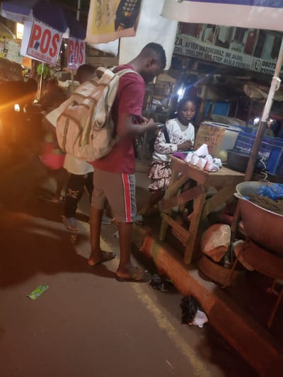 The image captures a bustling night market scene in Ibadan, Nigeria. In the foreground, a young man wearing a maroon shirt, grey shorts, and a light-colored backpack walks or stands with his bare feet visible, facing towards the right side of the frame. Behind him, a young woman in a light-colored top with a "Dior" logo is seated behind a wooden stall, looking towards the viewer.
The market features various wooden structures and stalls. A large reddish-brown basin filled with dark material sits on a wooden stand. Small white packages are visible on the woman's stall. The ground is dark, possibly asphalt, and contains some litter, including a green wrapper and a black plastic bag.
The scene is brightly lit by artificial light sources, creating strong glares and deep shadows, suggesting it is evening or night.
Visible text on signs provides commercial and location details: "ZA, AFTER MARDIEGRAZ, IRE AKARI, DHL, ORELOPE BUS STOP, AKALA EXPRESS" along with phone numbers "080738" and "0703716." Other signs declare "POS IS AVAILABLE HERE" and display "ALEX ARENAJU 2102958811," likely contact information for a vendor. The overall atmosphere is that of an active, informal street market.