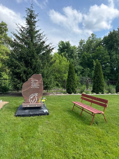 This is an eye-level, medium shot of a memorial located in a grassy area, likely a cemetery, in Chudovo, Russia. The memorial is a reddish-brown, irregularly shaped stone monument with inscriptions and a stylized flame design in white. At the base of the monument is a black rectangular pedestal. In front of the pedestal, on the grass, are artificial flowers. To the right of the memorial is a weathered red wooden bench. Behind the memorial and bench, a black metal fence separates the manicured lawn from a wooded area with tall evergreen and deciduous trees. The sky above is blue with scattered white clouds. The overall scene is serene and appears to be a place of remembrance.