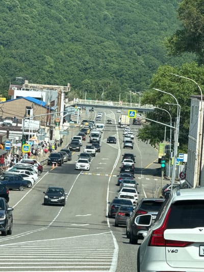 This image depicts a busy street scene in Lermontovo, Russia, characterized by significant vehicular traffic congestion. A multi-lane road stretches into the distance, lined with various commercial establishments and residential buildings on the left. A densely vegetated, steep hillside dominates the background.
On the right side of the road, a white building with vertical siding and a lighter-colored trim is prominently visible, along with utility poles and streetlights. A traffic light is illuminated green, indicating traffic flow. Several cars, including a white Volvo V60 in the foreground, are queuing in traffic.
The left side of the road features a pedestrian walkway with several people walking. Shops with awnings are present, and a bus stop sign is visible. A pedestrian overpass spans the road further in the distance. Cars are parked along the side of the road in some areas.
The scene is bathed in sunlight, suggesting daytime. The overall impression is one of a bustling, possibly tourist-oriented area with heavy reliance on road transportation.