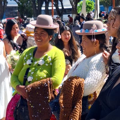 The image shows a group of people in what appears to be Cerro Colorado, Peru. The people are of Andean descent, wearing traditional clothing, including bowler hats with flowers. The woman in the foreground is wearing a bright green sweater adorned with white floral details. Some of the women are holding brown crochet bags. They are in a public space, possibly during a festival or cultural event, with a mix of trees and buildings in the background.