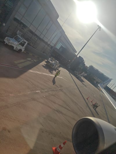 The image depicts an airport tarmac at Cologne Bonn Airport. A Ryanair airplane wing is visible in the foreground, with its engine cowling in the lower right corner. In the middle ground, a yellow safety vest-clad airport worker is walking on the tarmac, casting a shadow. Various ground support equipment, including a white baggage tractor and several orange traffic cones, are scattered across the tarmac. The airport terminal building, characterized by its modern glass facade, is visible on the left side of the image, with the gate designation "D02" clearly marked on its exterior. The bright sun is high in the sky, creating lens flare and casting long shadows. The scene is typical of an operational airport environment.