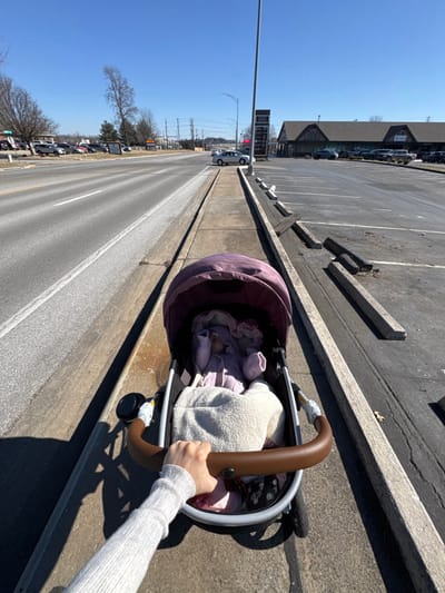 A baby stroller sits on a sidewalk, next to a multi-lane road on a sunny day. Inside, a baby is covered with purple clothes and a white blanket. A person in a gray sweater holds the stroller handle. The road runs parallel to a parking lot, which has several parked cars and a building in the distance. A light post stands tall near the road. The sky is blue and clear.