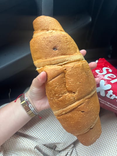 A person is holding a large croissant in their hand. In the background is a red package of "Ser...t" with writing in Greek. The person is wearing a bracelet with gold and silver accents. The setting appears to be inside a vehicle, indicated by the black interior. The patterned shirt of the person holding the croissant is visible. There is no evidence to confirm that this is the city of Μέλισσα, Greece.