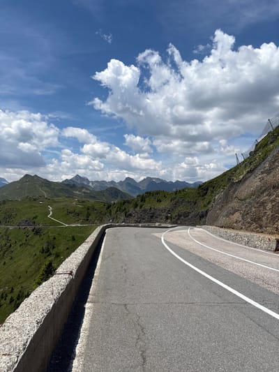 The image depicts a winding mountain road in the Italian Alps, likely near St. Leonhard in Passeier - San Leonardo in Passiria. The road curves sharply to the right, with a concrete barrier on the left and a steep, rocky embankment on the right. White lines mark the lanes of the asphalt road. Beyond the road, rolling green hills dotted with sparse trees stretch towards a majestic mountain range under a bright blue sky filled with white, puffy clouds. A mesh fence is visible on the embankment, designed to prevent rockfalls. The scene appears to be captured on a clear, sunny day. There are no people or vehicles visible in the image.