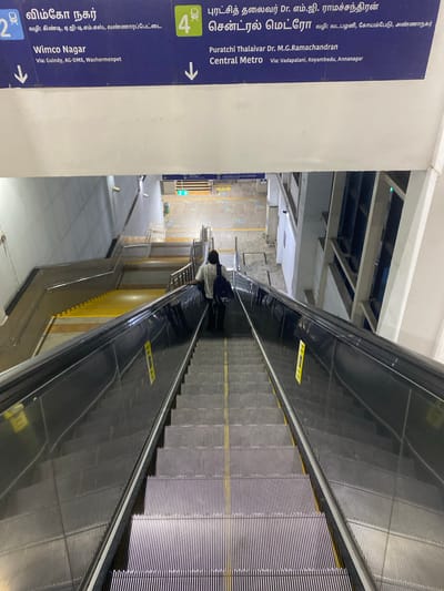 The image captures a scene inside a subway station, focusing on a downward-moving escalator. A person, partially visible, is ascending the adjacent escalator on the right side of the frame, facing away from the camera. The escalator steps are metallic and ribbed, with yellow accents along the sides. Above the escalators, a concrete ceiling with exposed beams is visible.

A prominent blue sign on the left displays directional information in both Tamil and English. It indicates "Wimco Nagar" and "Puratchi Thalaivar Dr. M.G. Ramachandran Central Metro" as destinations, with corresponding arrows pointing downwards. The Tamil text also provides route information, mentioning "Via: Guindy, AG-DMS, Washermenpet" and "Via: Vadapalani, Koyambedu, Annanagar." These signs suggest the location is within a public transportation system, likely a metro station in India, given the Tamil script and place names.

The overall lighting suggests an indoor environment, possibly during the day, with artificial lighting illuminating the station. The scene appears to be functional and orderly, typical of a transportation hub. The perspective is from an elevated position looking down at the escalators.