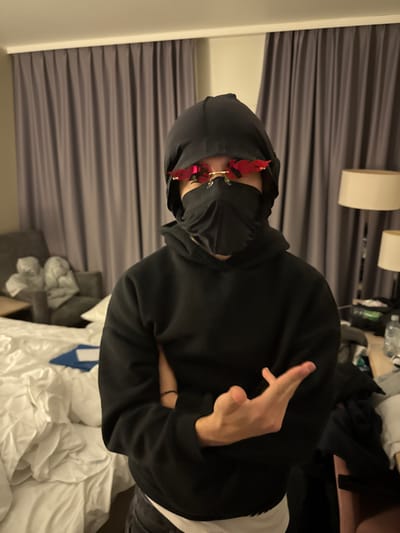 A person wearing a black hoodie, durag, face covering, and red flame-shaped sunglasses poses indoors. Their arms are crossed, and their right hand is raised, fingers extended in a gesture. The setting appears to be a hotel room with a bed, curtains, and lighting visible in the background. Clothes and bottles are strewn around the room.