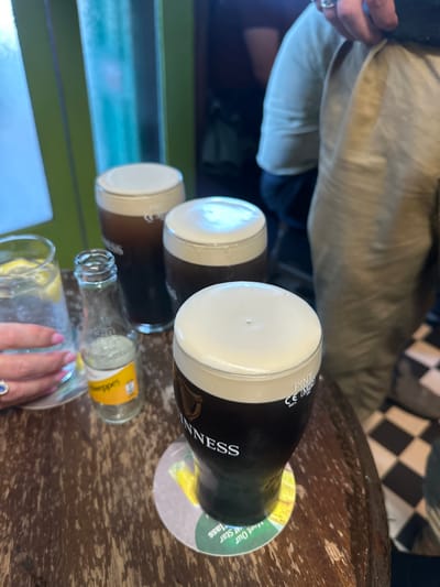 In the city of Westport, Ireland, on a weathered wooden table, sit three pints of Guinness, one with a prominent "Guinness" label. Next to them are a glass of water with lemon slices and a small, empty Schweppes bottle. In the background, a person's torso is visible, clad in a light-colored shirt and beige trousers, and another person's arm with a ring is extended towards the glass of water. The checkered floor suggests a pub or restaurant setting.