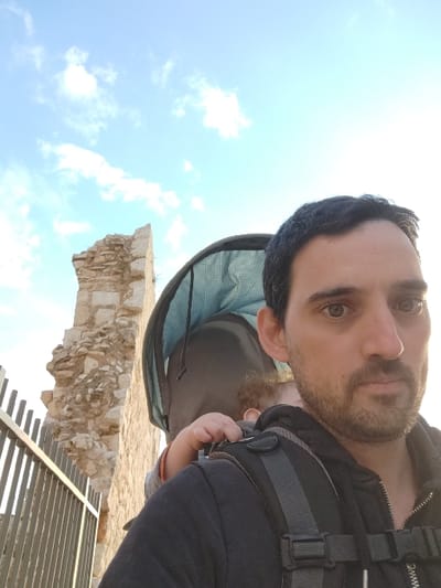 A close-up selfie captures a man and a child in a baby carrier outdoors on a bright day in Rosh HaAyin, Israel. The image, originally taken in portrait orientation, features the man prominently in the foreground, looking directly at the camera with a neutral expression. He has dark hair, a beard, and brown eyes, and is wearing a dark jacket or shirt. A robust baby carrier is strapped to his front, holding a partially visible infant or small child with curly hair and a tiny hand. The carrier's interior lining is a distinctive light blue or teal checkered fabric.

The background reveals light-colored stone ruins or natural rock formations, indicative of an archaeological or historical site, consistent with the Rosh HaAyin context. Above the ruins, the sky is a vibrant blue with scattered white clouds, suggesting clear weather. Strong, natural sunlight illuminates the scene, with a noticeable glare in the lower right, placing the time of day between late morning and early afternoon. In the upper left, a modern structure with horizontal slats, possibly a shaded walkway or viewing platform, can be seen. No text is visible in the media.