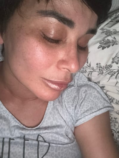 A close-up, overhead shot shows a person lying on a floral patterned bedspread. The person's face is in profile, with their eyes closed. They are wearing a light blue t-shirt with a subtle leopard print. The lighting in the image is warm and creates a subtle sheen on the person's skin. The image was taken in Bellmore, United States of America.