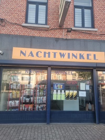 This is a vertical, eye-level, medium shot of the exterior of a "NACHTWINKEL" (night shop) in Heusden-Zolder, Belgium. The shop features a blue facade with a large display window filled with various products. The window showcases shelves stocked with bags of snacks, bottles of drinks, and other convenience items. A prominent orange sign with large blue lettering spells out "NACHTWINKEL". Inside the window, an "OPEN" sign with red neon lettering is visible, as is a notice stating "Dinsdag is rustdag" (Tuesday is rest day). Reflections in the glass reveal the street outside, including trees, buildings, and the sky. To the left of the shop, a brick building with two windows is visible. The ground in front of the shop is paved with grey bricks. The scene appears to be during daylight, with no specific indicators of weather conditions. The overall impression is that of a typical neighborhood convenience store.