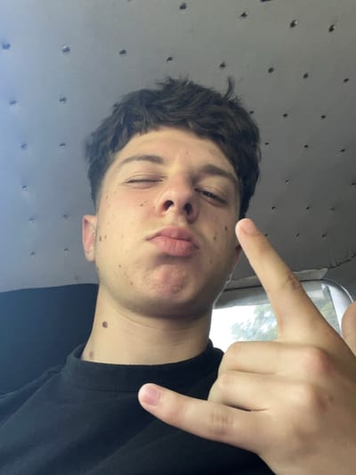 The image shows a close-up, selfie-style shot of a young person. The individual is inside a vehicle, evidenced by the textured grey headliner with visible circular fasteners in the upper portion of the frame, and the hint of greenery and sky visible through a window on the right. The person has dark, messy hair, and is making a kissing face with one eye closed. They are wearing a black t-shirt. Their right hand is raised, with the index finger and thumb extended to form a "rock on" gesture. The person has several moles visible on their face and neck. The lighting suggests it is daytime, possibly overcast due to the diffuse light. No specific landmarks of Taran, Bulgaria are visible.