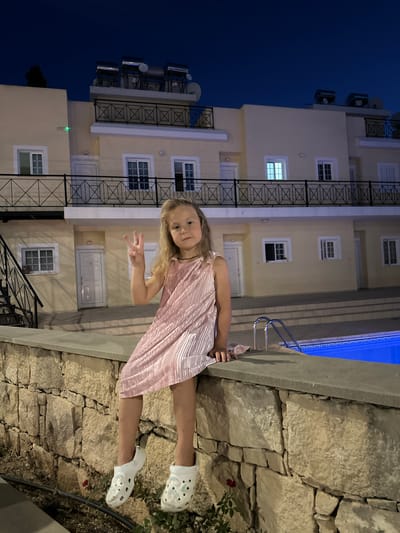 A young girl with blonde hair, wearing a pink pleated dress and white clogs, poses in front of a building at dusk. She is making a peace sign with her right hand and smiling at the camera. The building appears to be a hotel or apartment complex, with multiple windows and balconies. A swimming pool with bright blue water is visible to the right of the girl. The scene is lit by artificial lights, suggesting it is evening. The background shows a stone wall and some foliage. The overall atmosphere is relaxed and cheerful. The image appears to be taken in Peyia, Cyprus, as indicated by the location context.