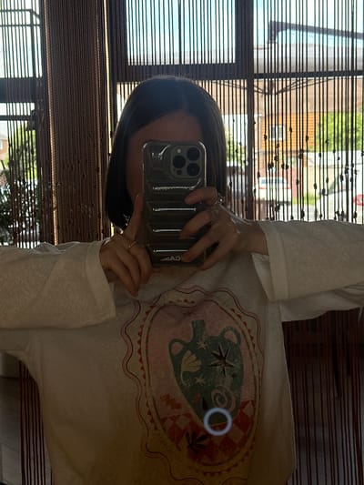 The image is a mirror selfie featuring an individual holding a dark olive-colored smartphone, which obscures their face. The person has dark, straight hair visible on either side of the phone. They are wearing a light-colored, possibly white or off-white, long-sleeved top with wide, flared cuffs. The front of the shirt displays a large, central graphic design in a stylized oval or shield shape. The graphic features an ornate jar or urn with handles, surrounded by abstract shapes, stars, and foliage motifs in muted tones of teal, pink, orange, and white, with a checkerboard pattern at the bottom.
Rings are visible on the fingers of both hands; the right hand wears a thin band and a wider, textured band, while the left hand has a single thin band. The smartphone case, partially showing "CASETiFY" branding in reverse, features a prominent triple-lens camera array.
The background, reflected behind the individual, consists of an interior space with a window covered by vertical dark brown bead curtains or blinds. Through these translucent blinds, an outdoor scene is visible. This exterior view includes a bright sky, green trees and foliage, and architectural elements of distant buildings, such as light-colored walls and parts of a roofline. The scene is illuminated by natural daylight. No specific geographical identifiers for Усады, Russia, are discernible within the image.