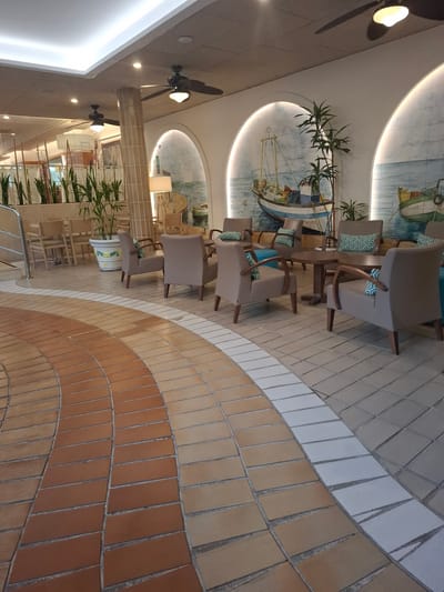 The image depicts an indoor seating area with a curved tile floor. The floor is laid with rectangular terracotta tiles arranged in concentric arcs. A white tile border separates the terracotta tiles from a lighter colored tile floor in the background. In the background, there are several upholstered chairs and round tables arranged in a lounge-like setting. The chairs are light brown and feature decorative green and white patterned cushions. Behind the seating area, there are arched murals on the wall depicting scenes of fishing boats on the water. Two ceiling fans are visible, one on the left and one on the right, both dark colored. The lighting appears to be a mix of recessed ceiling lights and decorative pendant lights. There are also several potted plants placed around the seating area, adding a touch of greenery to the space. The overall impression is one of a relaxed and well-lit indoor space, possibly a hotel lobby, cafe, or restaurant. The location is identified as Salou, Spain.