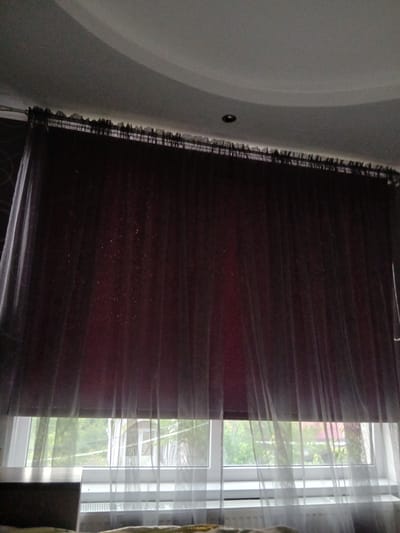 The image depicts a window in a room, likely a bedroom, in Vita-Poshtova, Ukraine. The window is covered by a dual-layer curtain system. The inner layer appears to be a dark-colored roller blind, possibly with a textured or slightly iridescent finish, that is partially lowered. Overlying this is a sheer, translucent white or light-colored curtain, which drapes from a rod at the top. This sheer curtain has a ruffled or gathered edge along the top. Through the window, a blurred view of greenery and trees is visible, suggesting an outdoor environment with vegetation. Below the window, a radiator is partially visible, a common fixture in Ukrainian homes. The ceiling of the room is visible at the top of the image, with a curved design. The lighting appears to be ambient, possibly from the outdoors or interior lighting that is not directly visible. No people or specific actions are depicted.