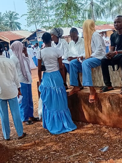 A group of students, predominantly in light blue and white uniforms, are gathered outside. Some wear head coverings of various colors. They appear to be standing or sitting near a building with a corrugated roof, with trees visible in the background. The ground is covered with leaves and debris.